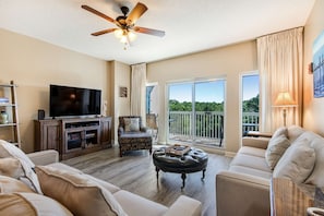A spacious living room with a flat-screen TV, a ceiling fan, a large window with a view, and a balcony. - Exclusive 2BR condo with beach access (Miramar Beach)
