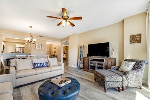 A living room with a sofa, ottoman, and armchair, a ceiling fan, a flat-screen TV, and a kitchen in the background. - Exclusive 2BR condo with beach access (Miramar Beach)