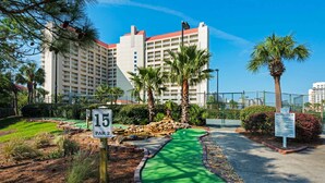 A golf course with a green putting area, a sign indicating hole 15, and a building in the background. - 2BR condo with pool view & private balcony (Miramar Beach)
