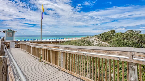 A wooden boardwalk leading to a beach with a flagpole, a lifeguard tower, and beach umbrellas. - 2BR condo with pool view & private balcony (Miramar Beach)
