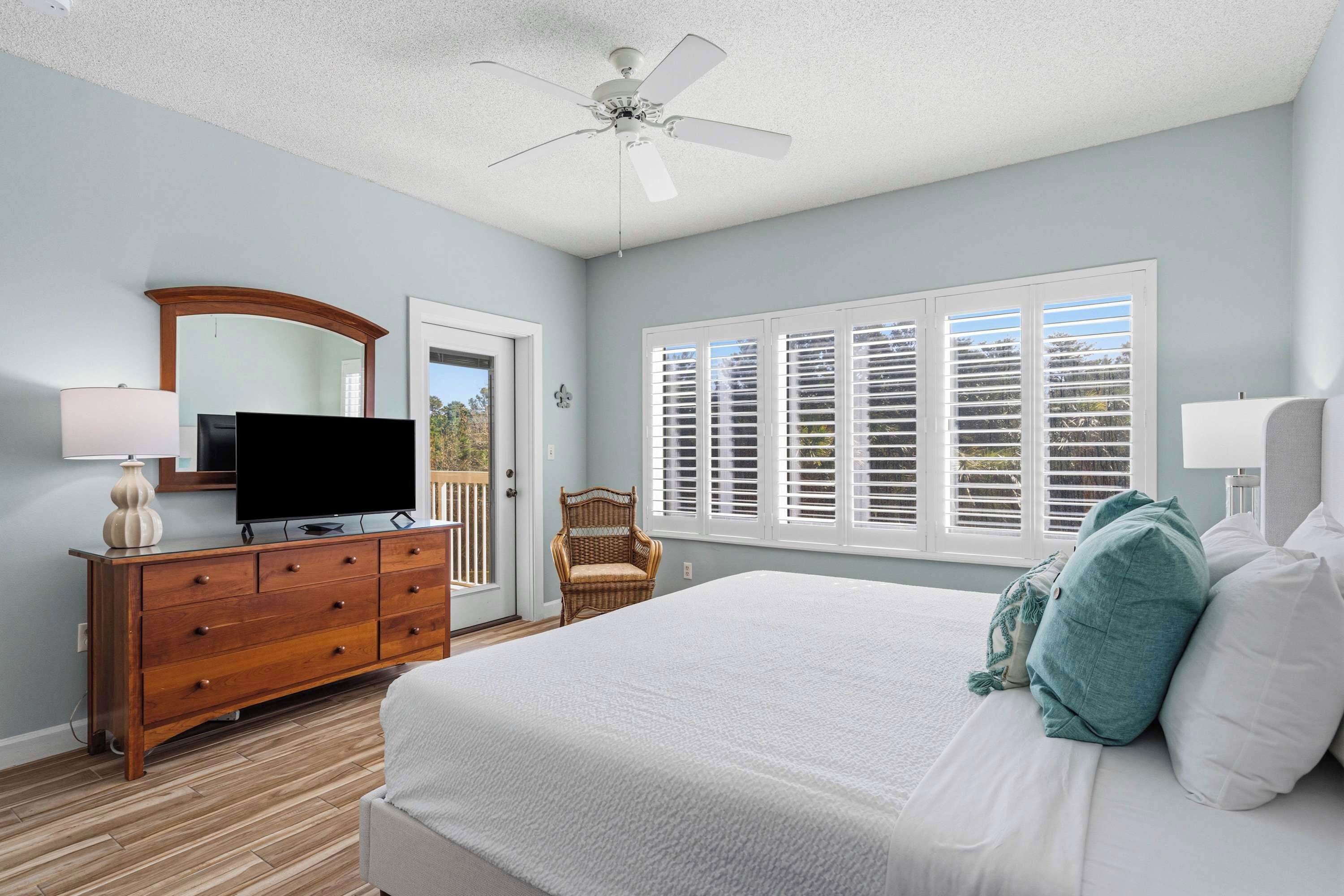 A bedroom with a bed, a dresser, a television, a ceiling fan, and a window with shutters.