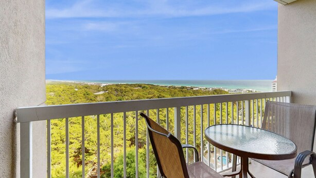 Ein Balkon mit Tisch und Stühlen, der auf eine Küstenlandschaft mit viel Grün und Blick aufs Meer blickt.