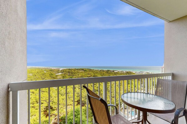 A balcony with a table and chairs, overlooking a coastal landscape with greenery and a view of the sea. - 1BR retreat: hot tub, steps to beach access (Miramar Beach)