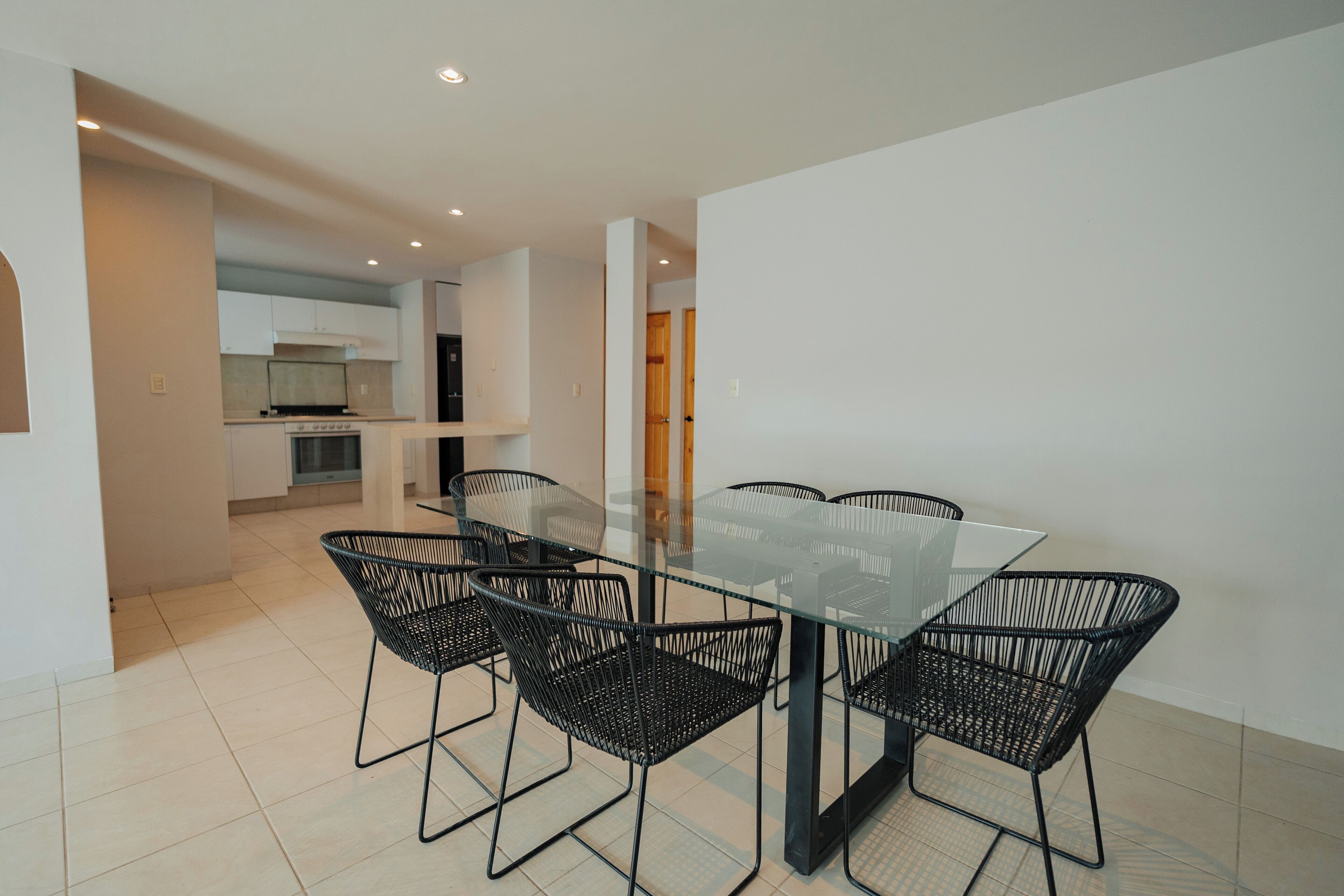 A modern dining area with a glass table and black wireframe chairs, leading to a kitchen with white cabinets and a stove.