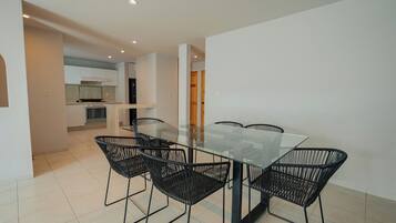 A modern dining area with a glass table and black wireframe chairs, leading to a kitchen with white cabinets and a stove.