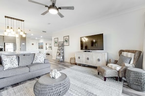 Een moderne woonkamer met een grijze bank, een gestreept voetenbankje en een rieten fauteuil. Een plafondventilator en een aan de muur bevestigde televisie zijn zichtbaar.