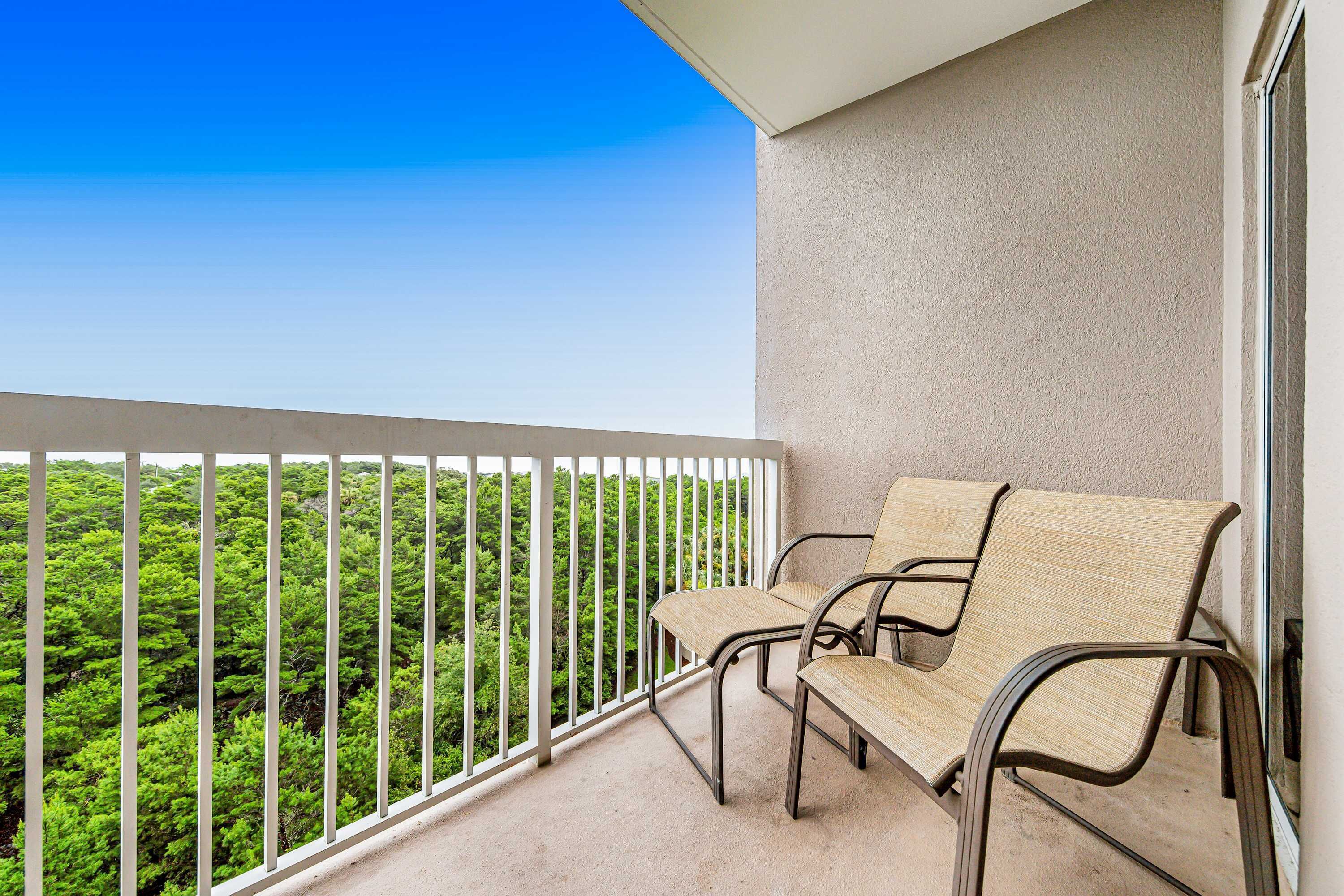 A balcony with two wicker chairs, a white railing, and a view of dense green foliage.