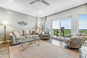 Ein Wohnzimmer mit Sofa, Sessel, Glastisch und Ausblick ins Freie.