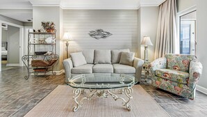 Un salon avec une table basse en verre, un canapé gris, un fauteuil à motifs floraux et un élément décoratif mural.