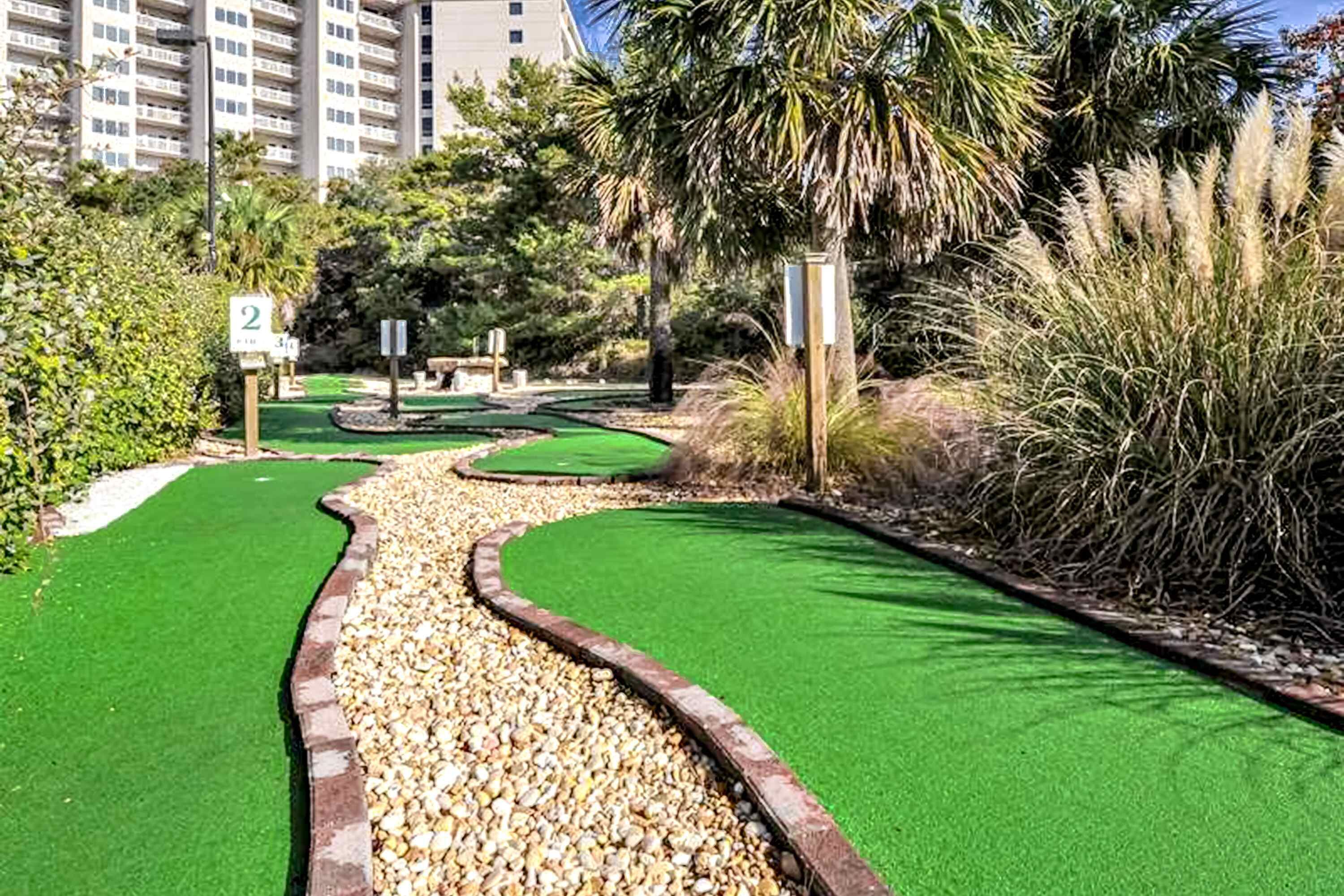A golf course with a green path, palm trees, and a building in the background.