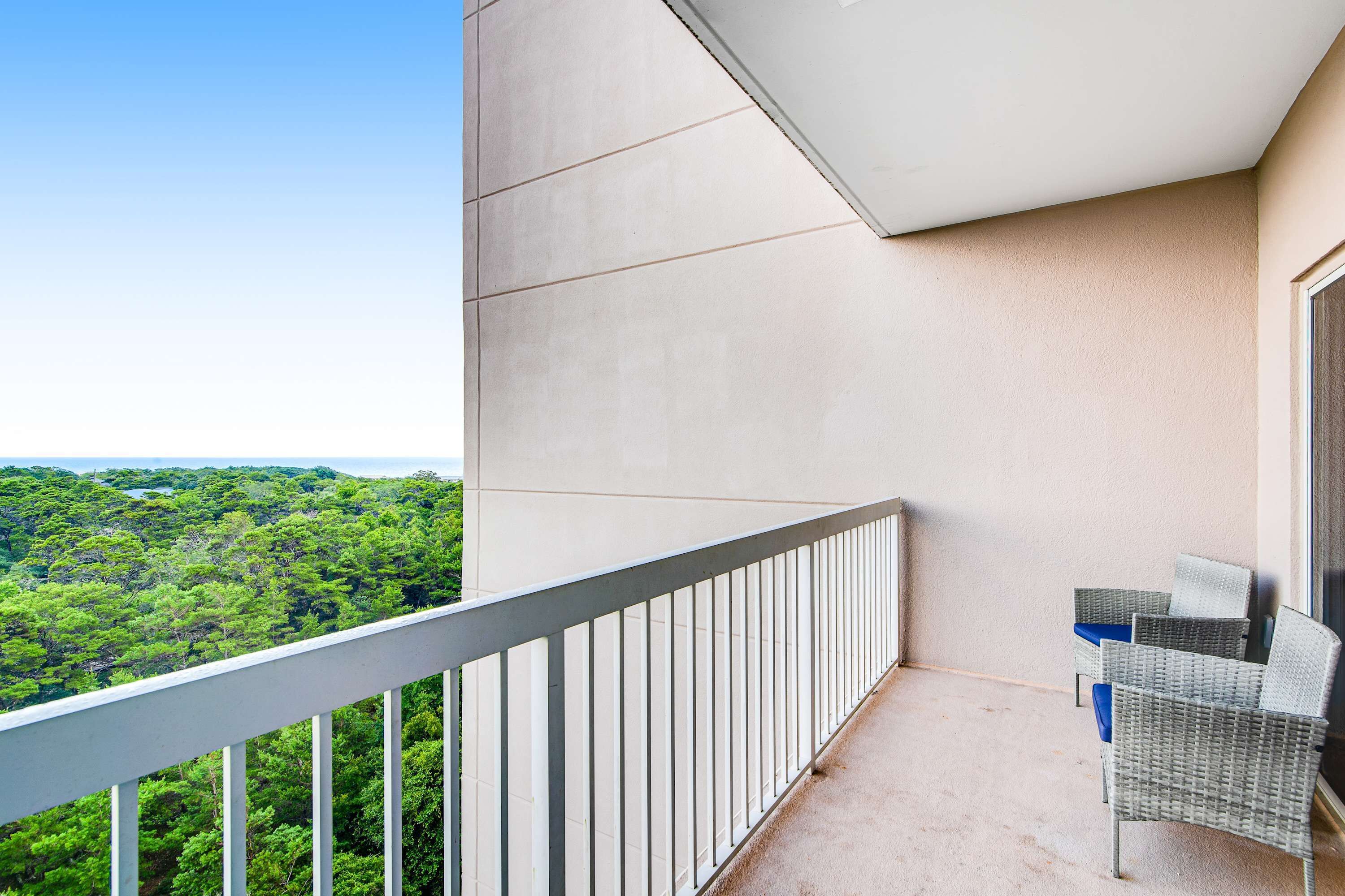 A balcony with a view of a forested area, a white railing, and two wicker chairs.