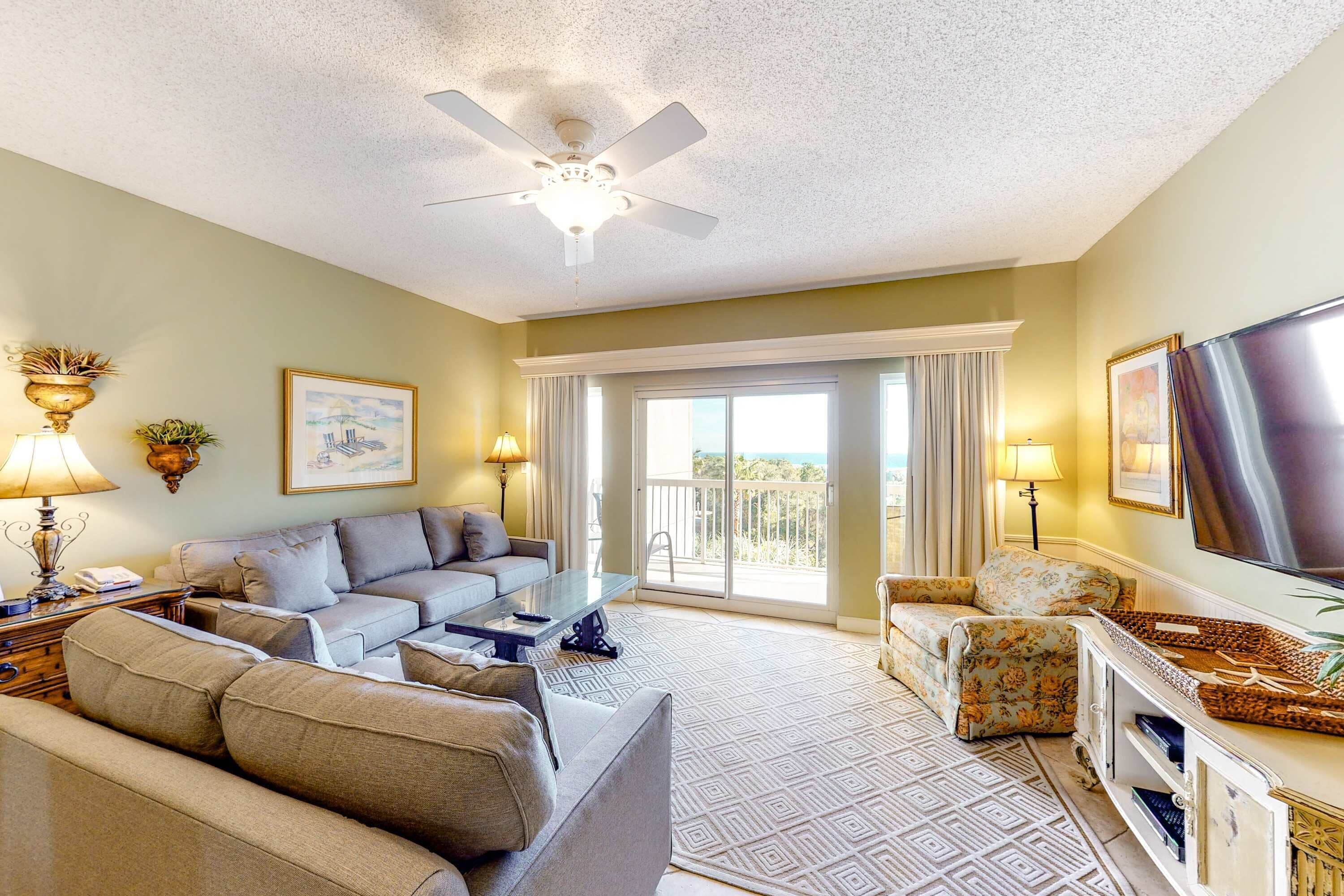 A living room with a ceiling fan, a sofa set, a coffee table, a TV, and a balcony with curtains.