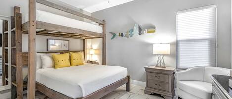 A bedroom with a wooden bunk bed, bedside tables, a white chair, a ceiling fan, and a window with blinds.