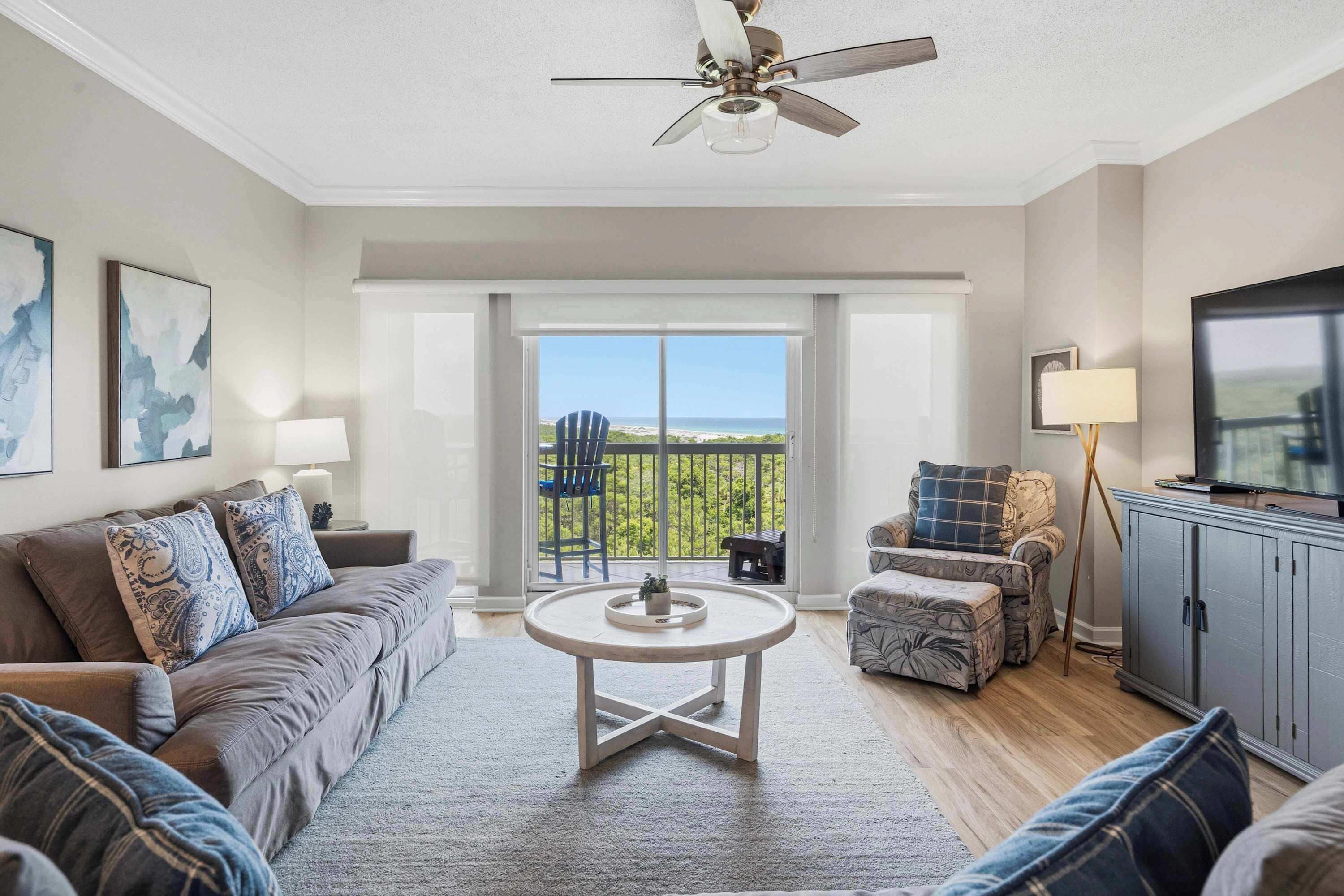 Un salon comprenant un canapé, un pouf, une table basse ronde, un ventilateur de plafond et une vue sur l’extérieur, accessible par une porte-fenêtre coulissante.