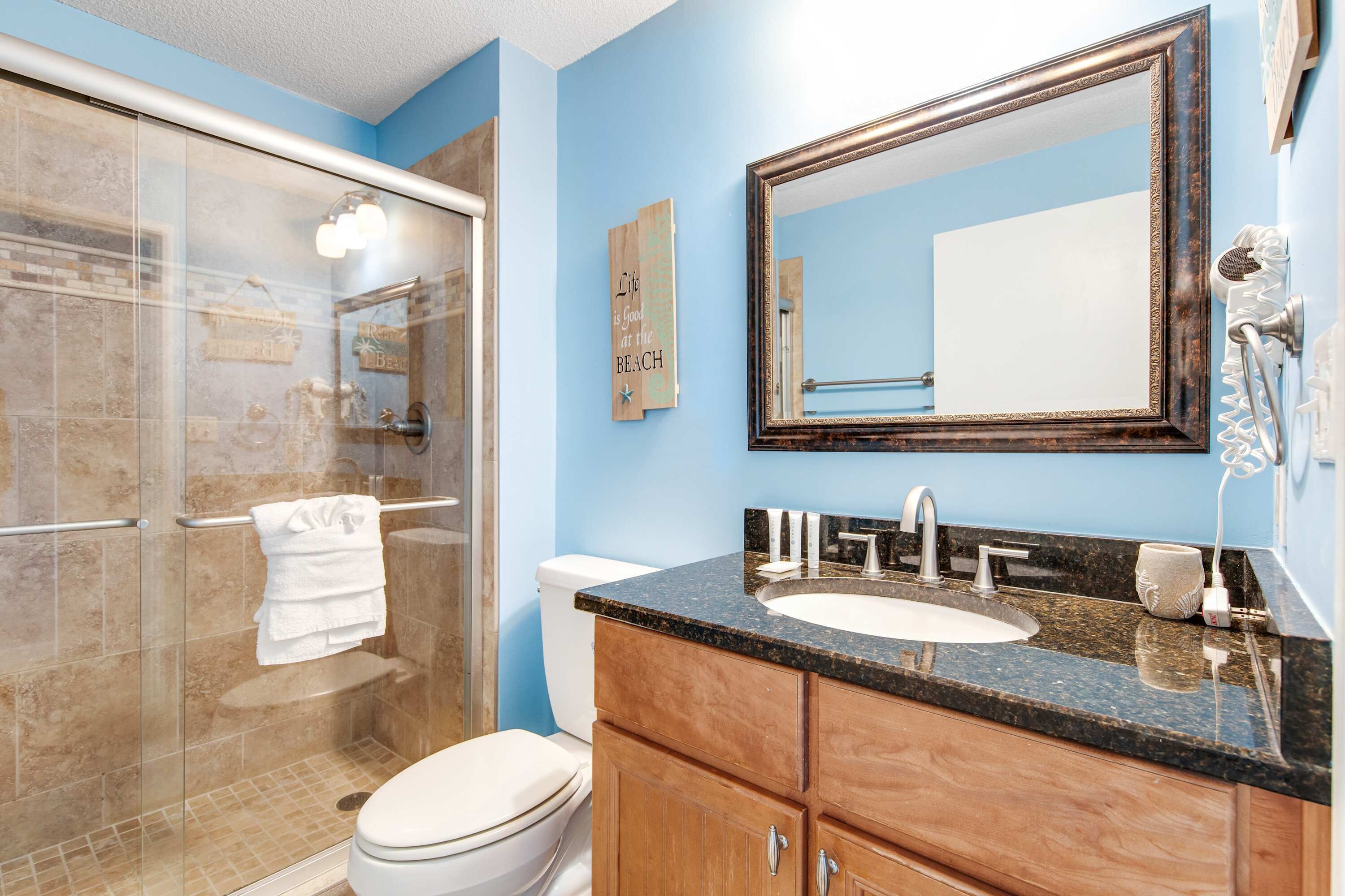 A bathroom with a glass shower enclosure, a white toilet, a sink with a black countertop, and a large mirror above the sink.