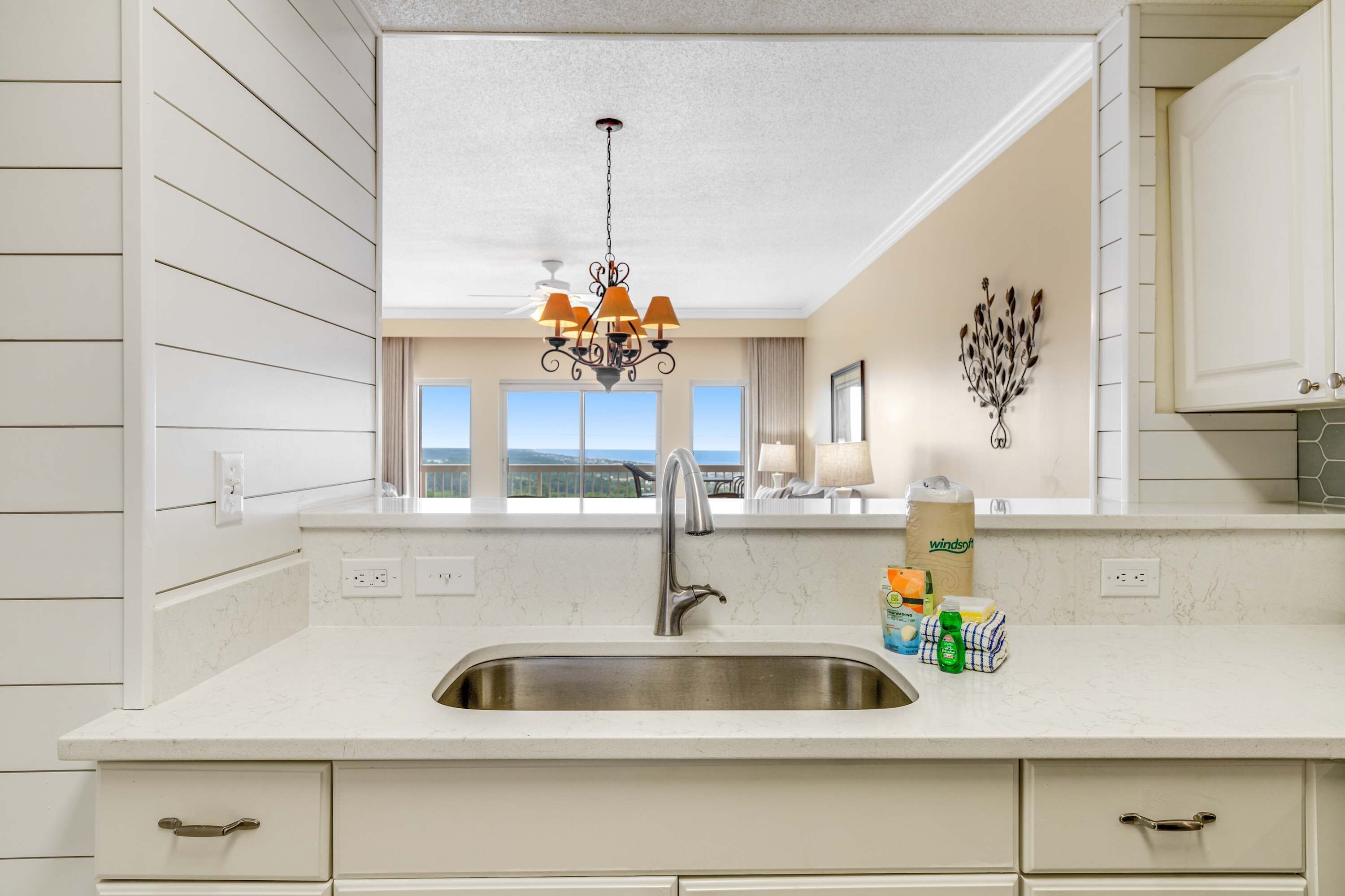 Eine Küche mit Spüle aus Edelstahl, weißen Schränken, einer Chandelüster und Blick durch das Fenster.
