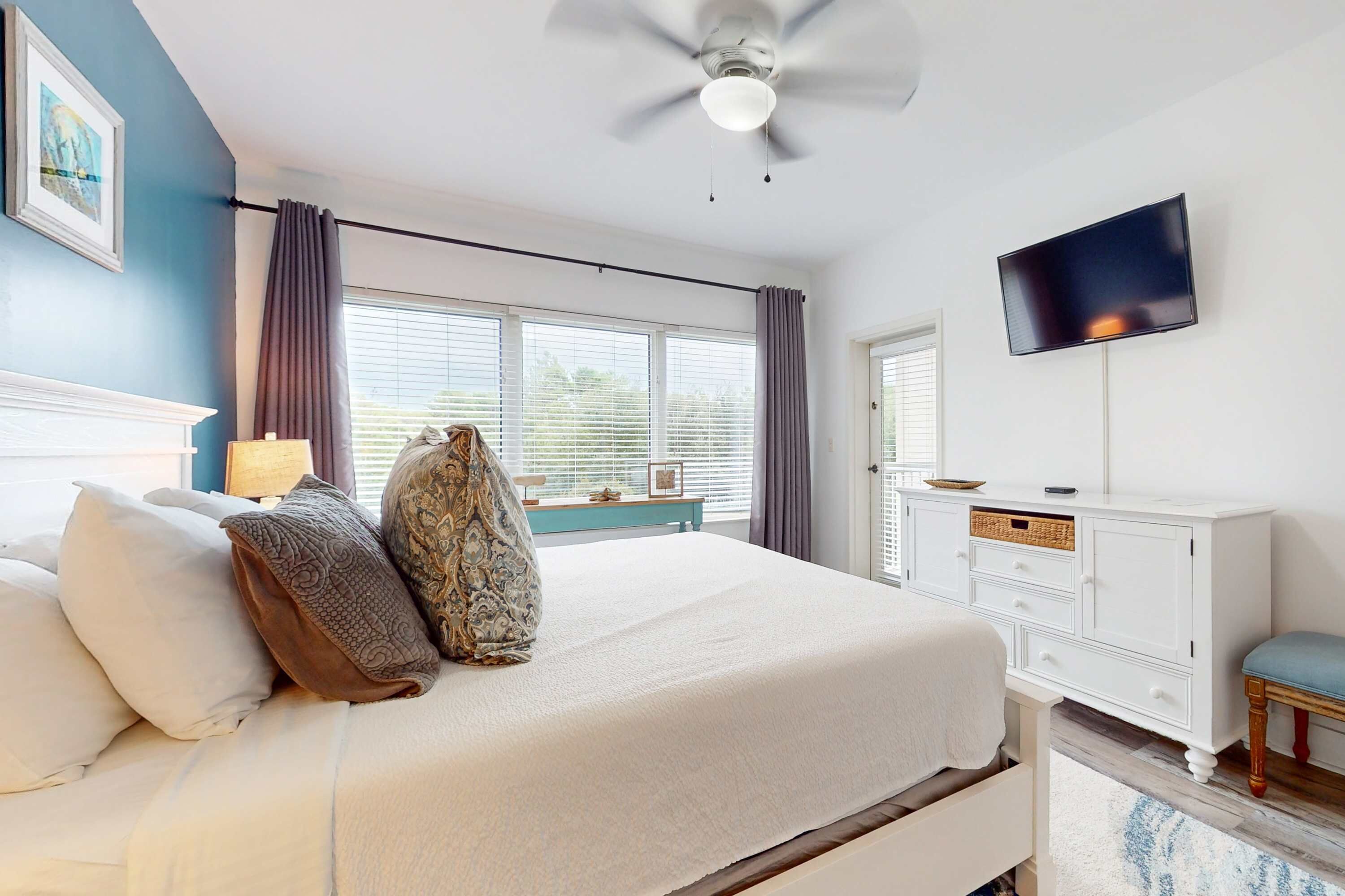 A bedroom with a bed, a ceiling fan, a TV, a window with curtains, and a white dresser.