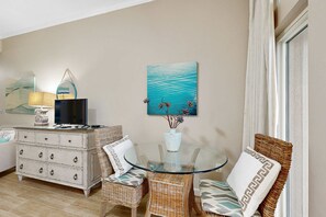 Ein Wohnzimmer mit Glastisch, Korbstühlen, einer weißen Kommode und einem Gemälde von einem blauen Meer.