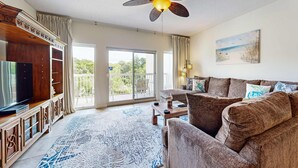 Una sala de estar con ventilador de techo, televisor, sofá, mesa de centro y un ventanal con vistas a zonas verdes.