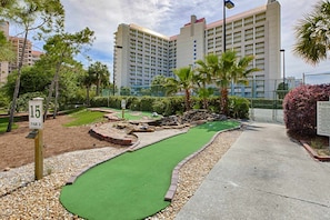 A miniature golf course with a green putting surface, surrounded by a gravel path and landscaped areas, with a large hotel building in the background. - Beachside studio with pool & hot tub access (Miramar Beach)
