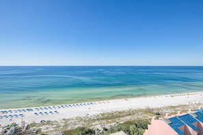 A beach view with clear blue water, a sandy shore, and a building with a blue roof. - Beachside studio with pool & hot tub access (Miramar Beach)