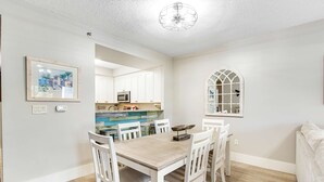 Zona pranzo con tavolo in legno e sedie bianche, cucina sullo sfondo e una finestra decorativa.