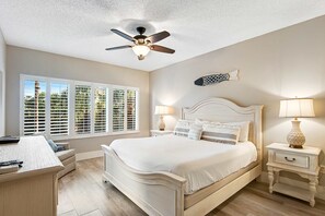 Ein Schlafzimmer mit Bett, Nachttischen, Deckenventilator, Fenster mit Jalousien und Blick auf Grünfläche.