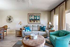 A living room with a beige sofa, a teal armchair, a round ottoman, a coffee table, a painting, and a decorative wall piece. - 2BR condo with Gulf views, pools, tennis (Miramar Beach)