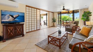 A spacious living room with a television, a sofa, and a coffee table. There is a sliding door leading to a balcony with a view of greenery.