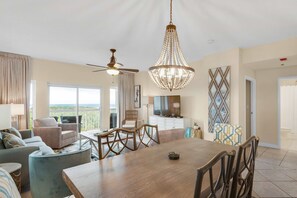 A spacious living area with a dining table, chairs, a chandelier, a ceiling fan, and a television. - 3BR Gulf View Condo w/ Hot Tub Access (Miramar Beach)