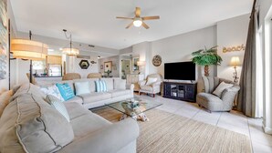 A living room with a sofa, armchairs, a glass coffee table, a TV, a ceiling fan, and a plant. - Exclusive Gulf View 2BR with Pool Access (Miramar Beach)