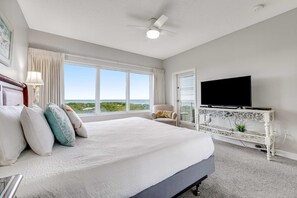 A bedroom with a large bed, a television on a white console, a ceiling fan, and a view of the outdoors. - Exclusive Gulf View 2BR with Pool Access (Miramar Beach)