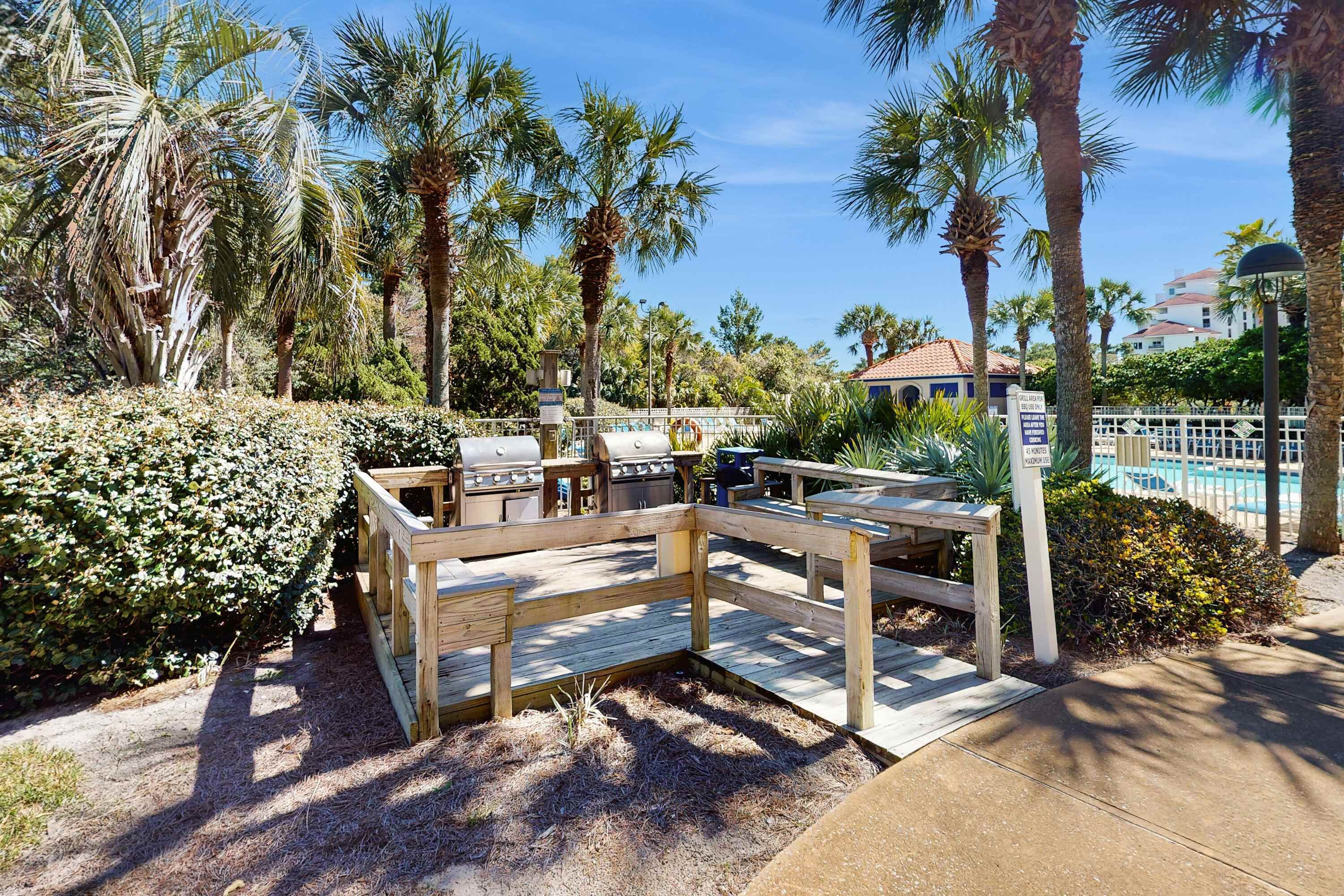 A wooden deck with barbecue grills, surrounded by palm trees and a pool.