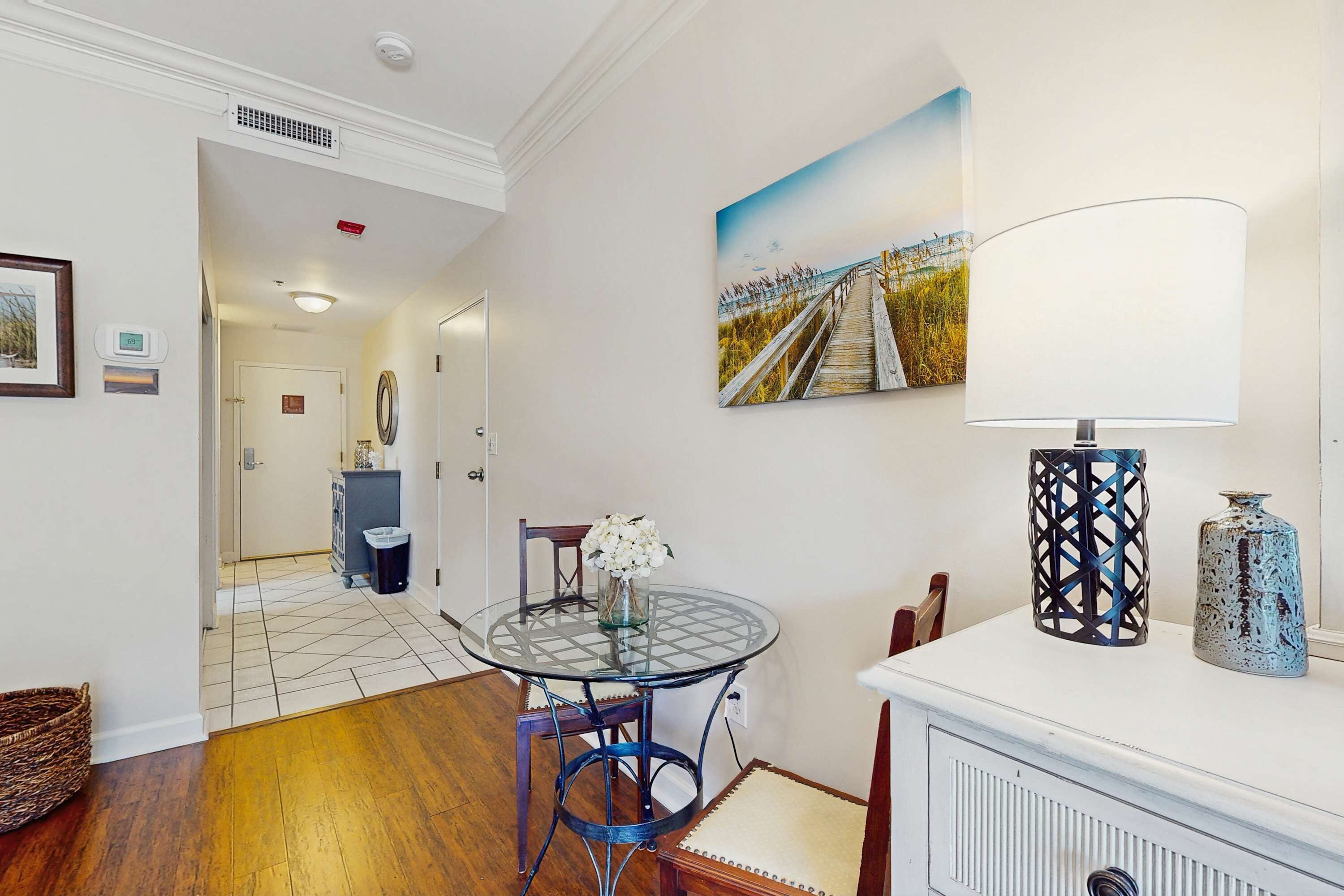 A dining area with a glass table, chairs, a white cabinet, and a painting of a beach boardwalk.