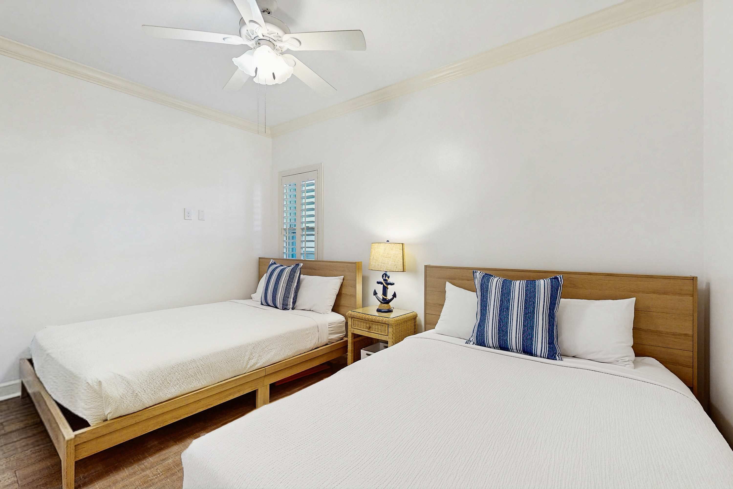 A hotel room with two beds, a ceiling fan, a nightstand with a lamp, and a window with blinds.