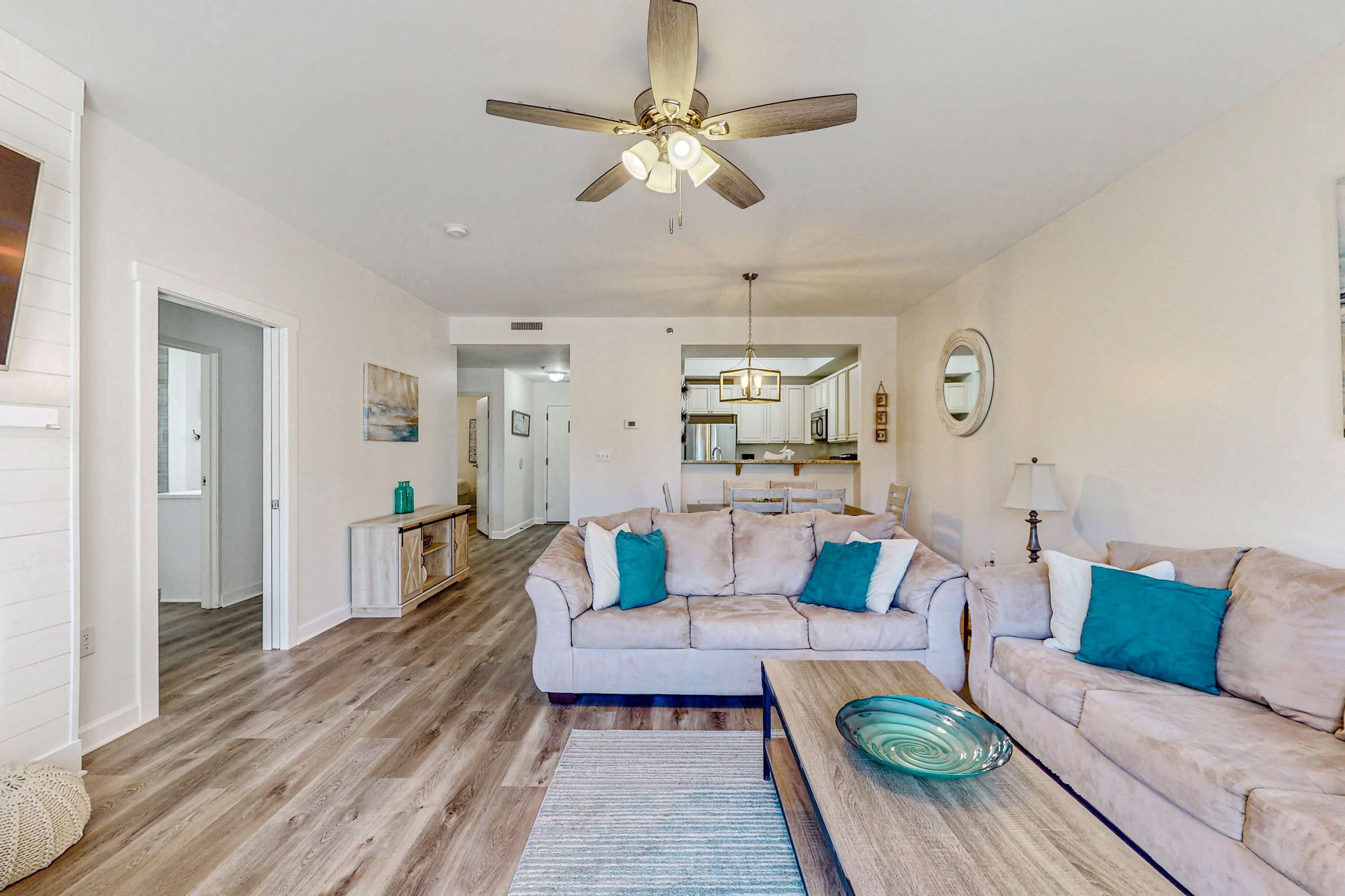 A spacious living room with a ceiling fan, a sofa set, a coffee table, and a kitchen area in the background.