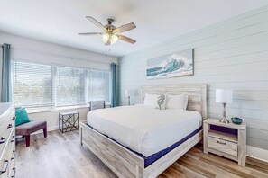 A bedroom with a wooden bed, a ceiling fan, a window with blinds, a nightstand, and a wall-mounted painting. - 2BR Resort Condo: Balcony, Pool, Beach Access (Miramar Beach)