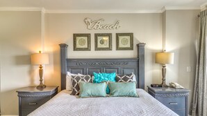 A neatly made bed with a quilted cover, two bedside tables with lamps, a wall-mounted artwork with the word 'Beach,' and a decorative headboard. - Scenic balcony views & resort pool access (Miramar Beach)