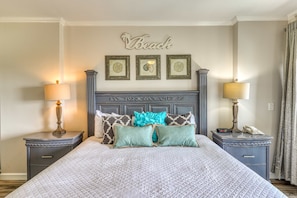 A neatly made bed with a quilted cover, two bedside tables with lamps, a wall-mounted artwork with the word 'Beach,' and a decorative headboard. - Scenic balcony views & resort pool access (Miramar Beach)