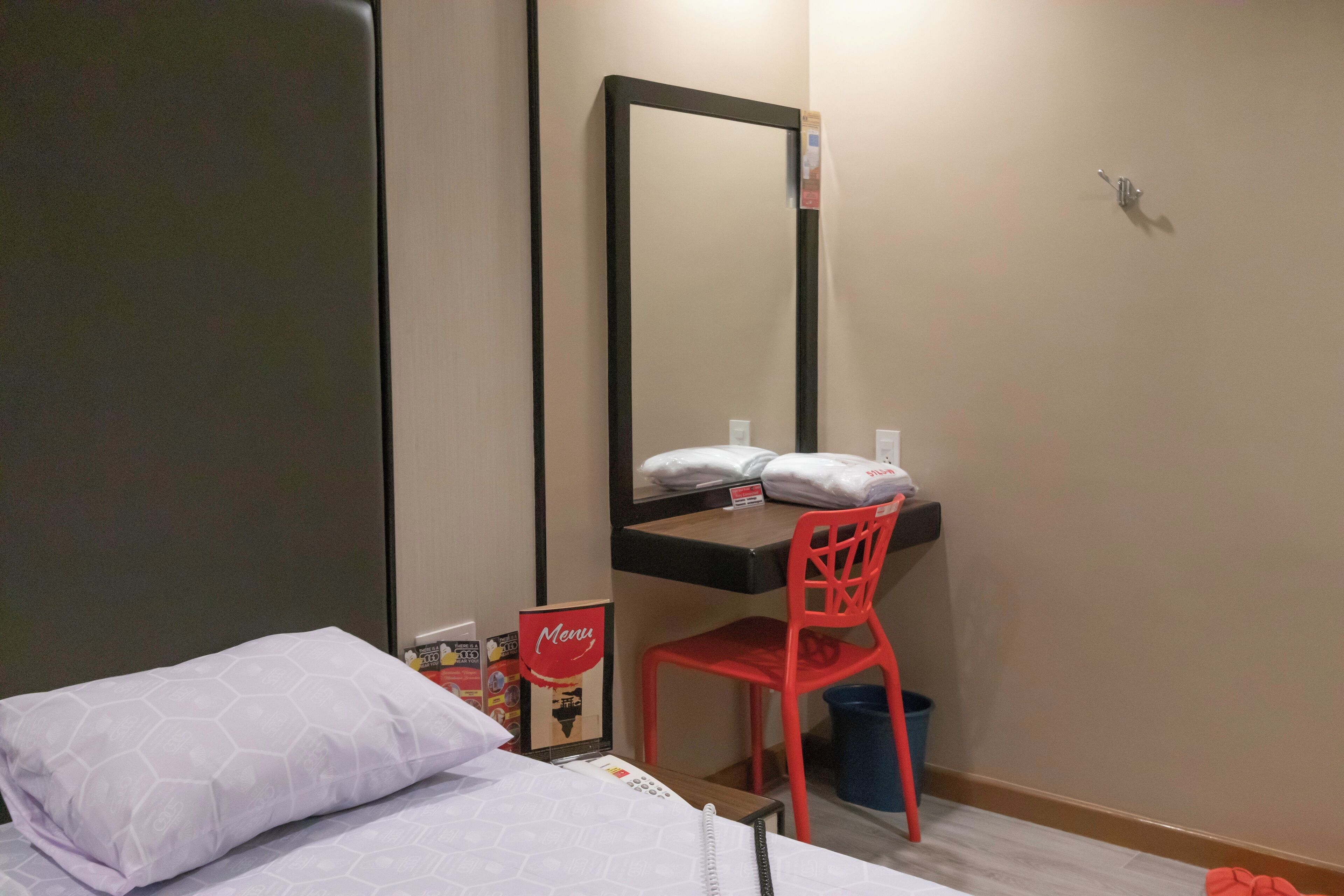 Une chambre d’hôtel avec un lit, un bureau doté d’un miroir, une chaise rouge et une petite poubelle.