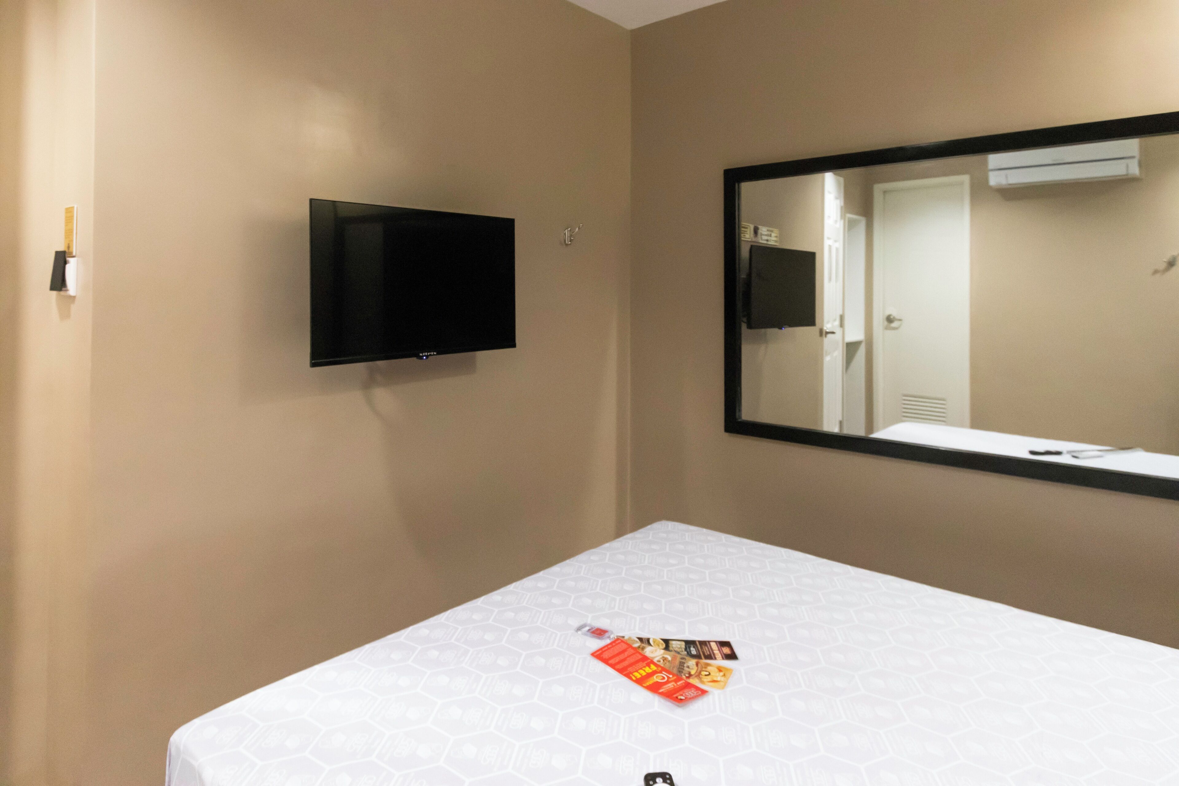 Quarto de hotel com cama, televisão montada e um espelho grande refletindo uma porta e um ar condicionado.