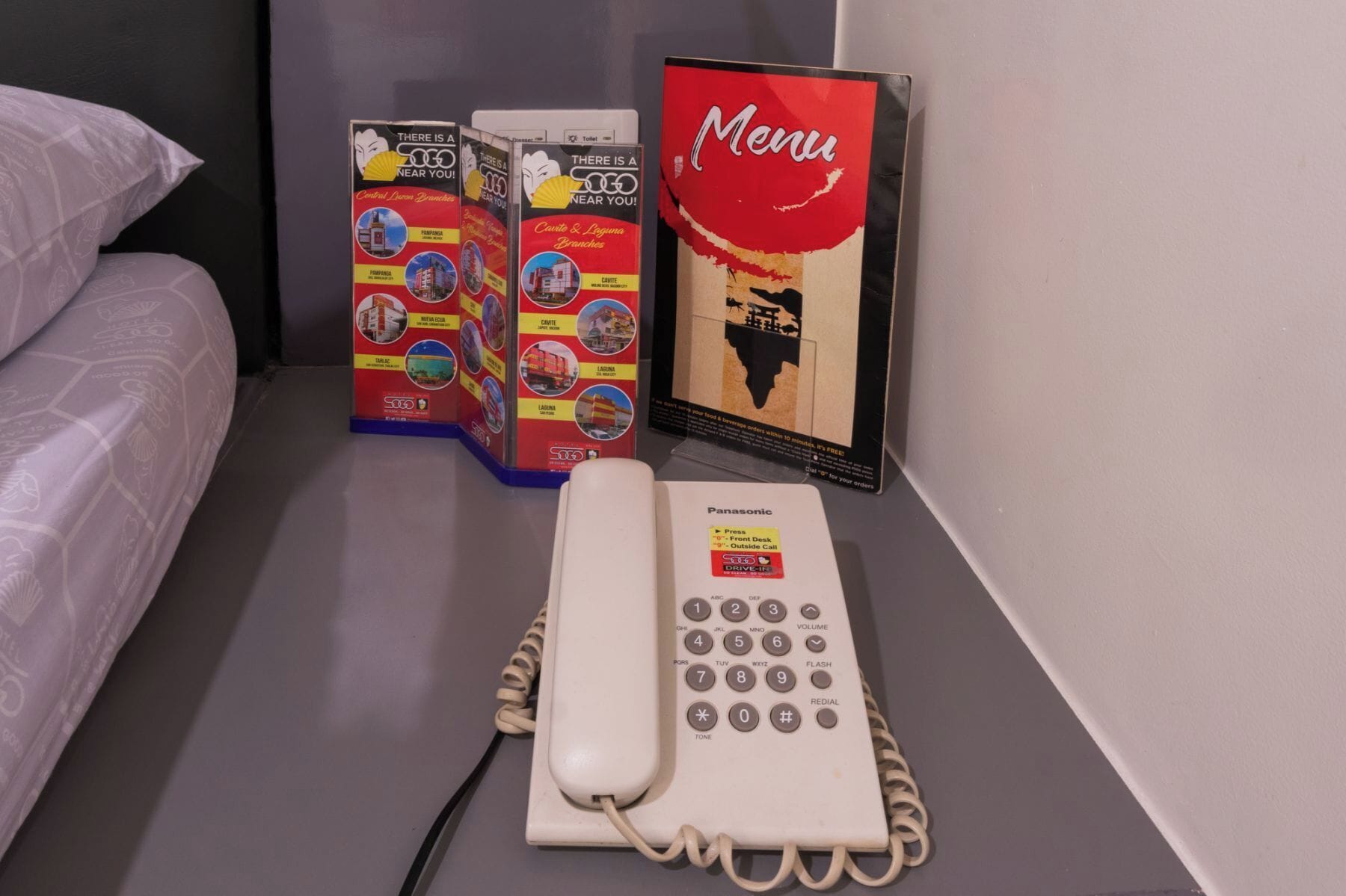 Une chambre d’hôtel avec un lit, un téléphone Panasonic et des brochures d’information sur la table de chevet.