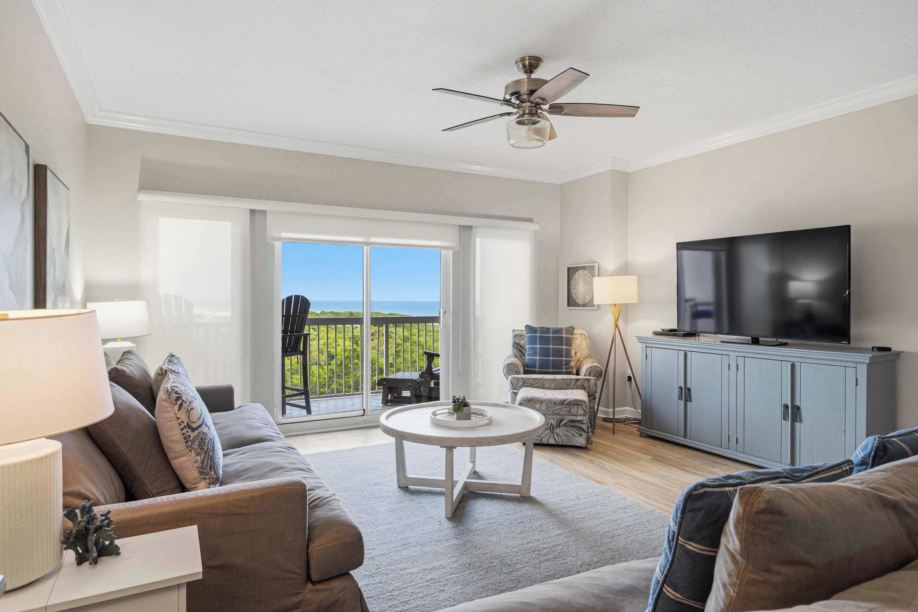 A living room with a sofa, a coffee table, a television, and a balcony with a view.