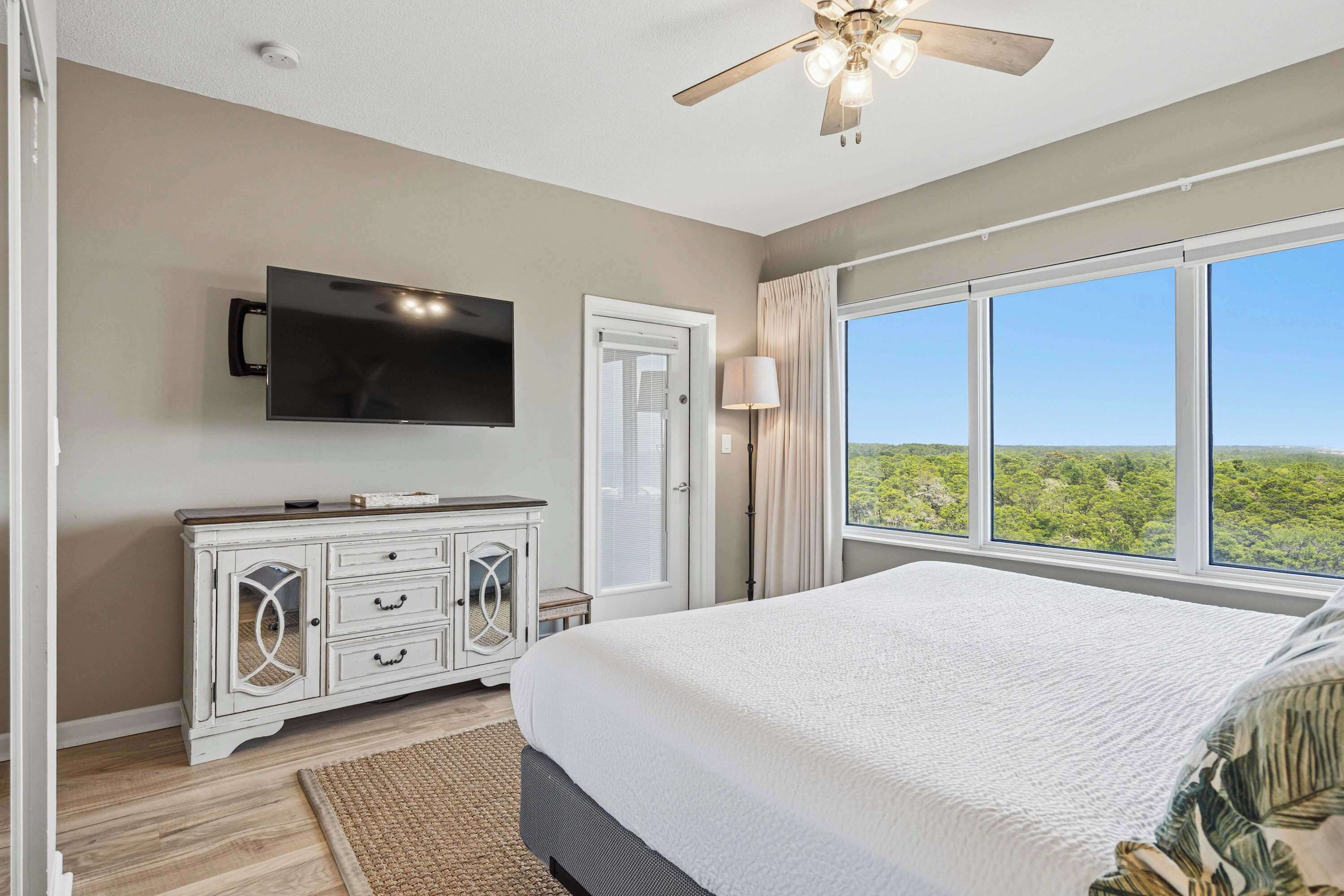 A bedroom with a bed, a television, a ceiling fan, and a large window with a view of greenery.