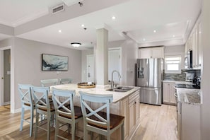 A modern kitchen with a central island, stainless steel appliances, and a dining area with light blue chairs. - Exclusive Gulf View Condo, Sleeps 5 (Miramar Beach)