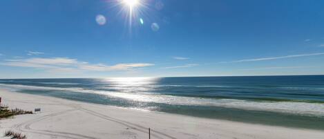 A beach with a clear sky, a bright sun, and calm ocean waves.