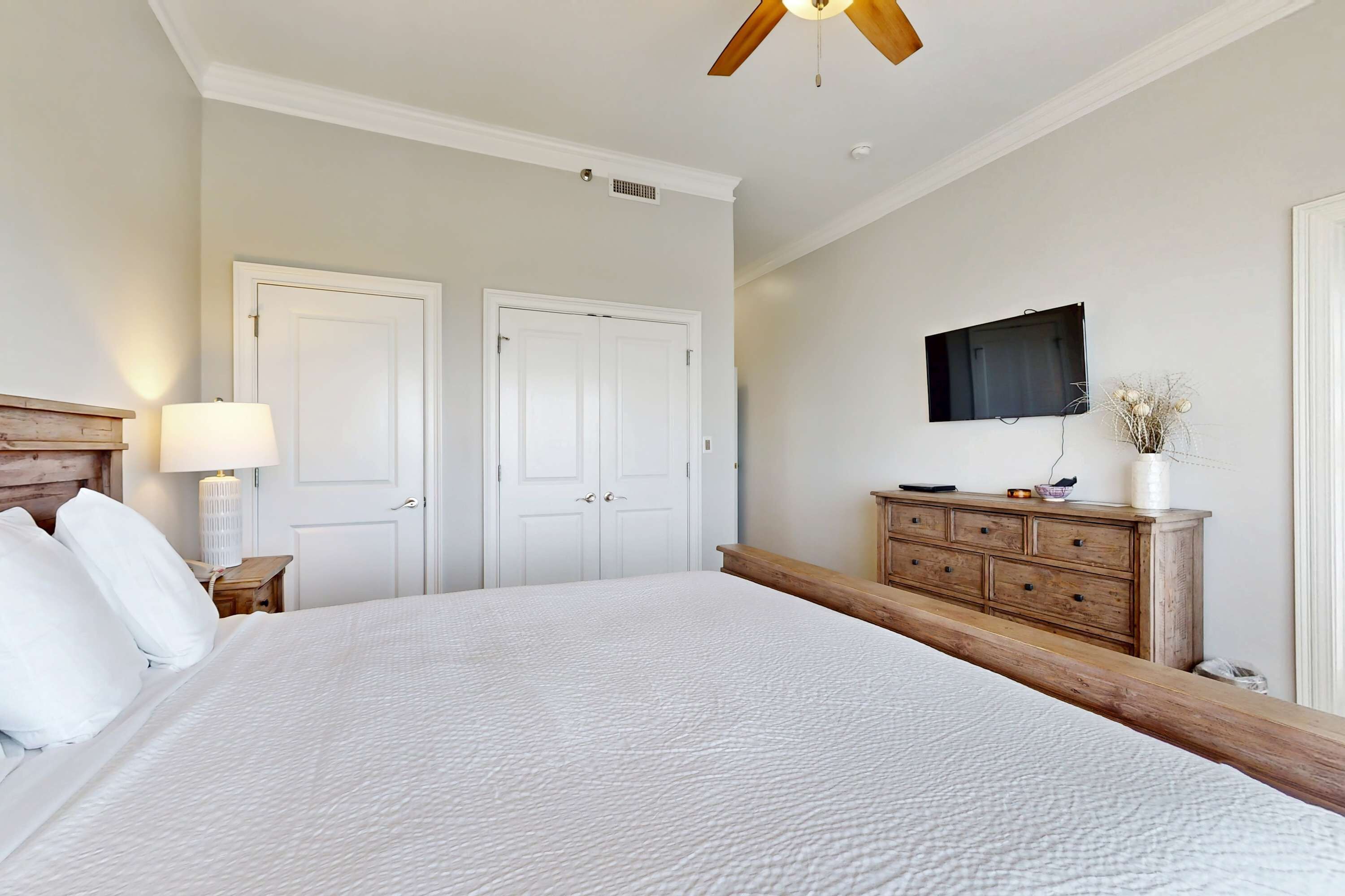 Une chambre avec un grand lit, deux portes blanches, une commode en bois, un téléviseur fixé au mur et un ventilateur de plafond.