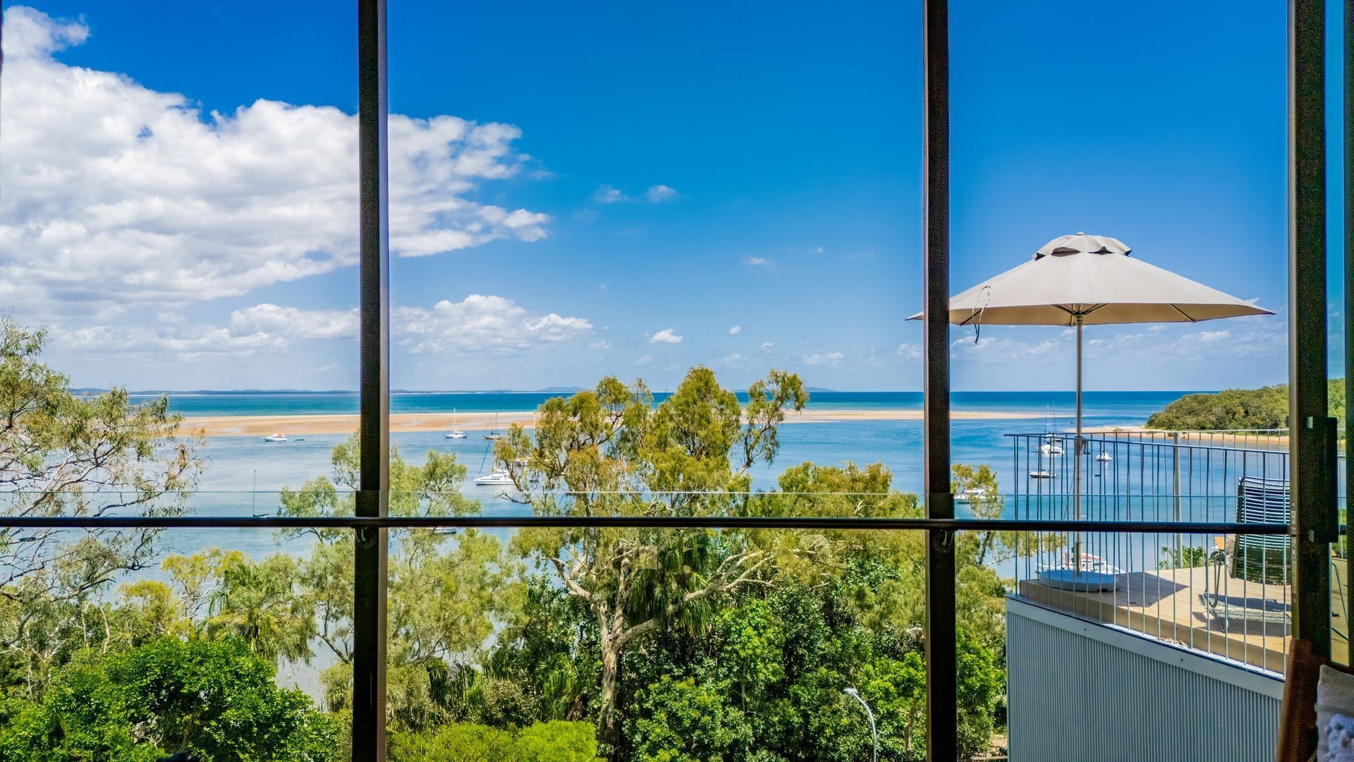 A view from a balcony with a beach and ocean in the background, a large umbrella, and lush greenery.