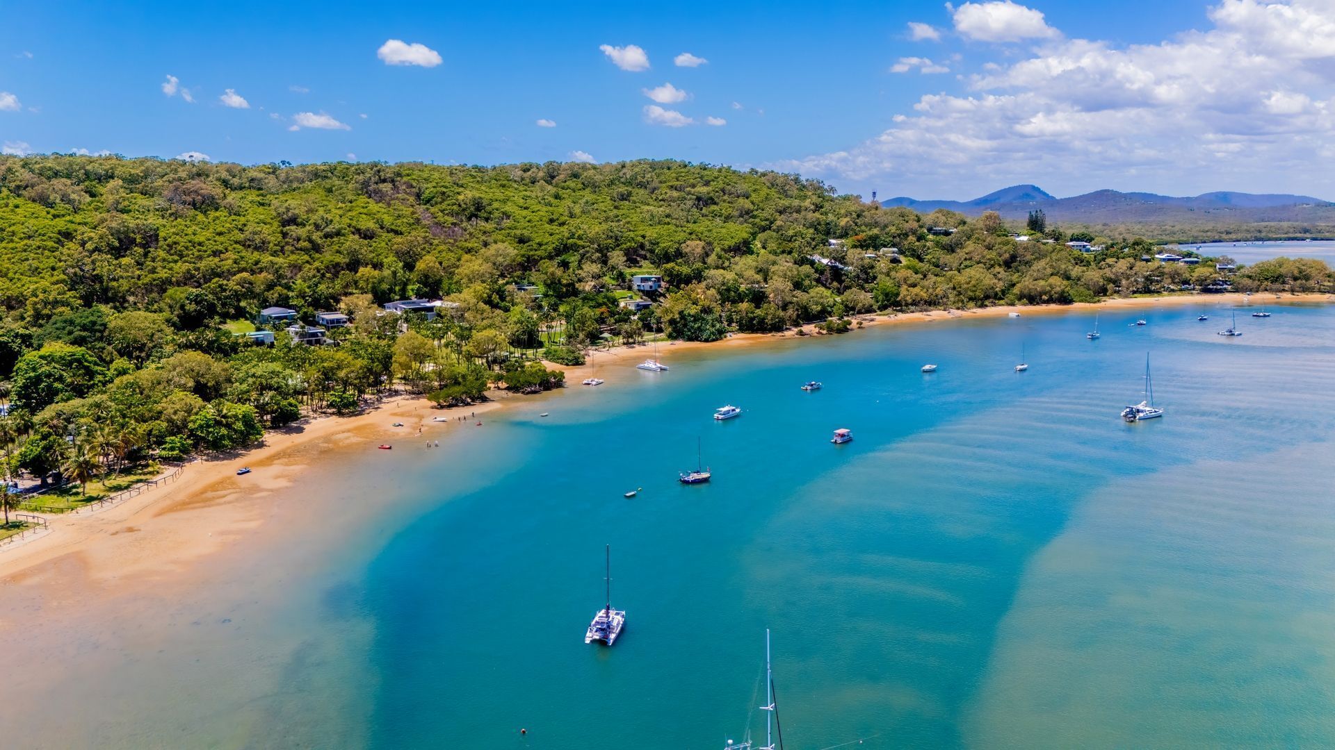 An aerial view of a coastal area with clear turquoise waters, a sandy beach, and a dense forested hillside. Several boats are anchored in the water, and there are buildings along the shore.