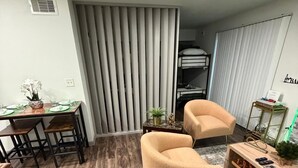 A modern living room with a wooden floor, a dining area with a table and chairs, two armchairs, a small side table with a lamp, and a bunk bed in the background. - Modern Studio Apartment Pool & Gym Access Ideal for Relaxing Stays in Houston (Houston)